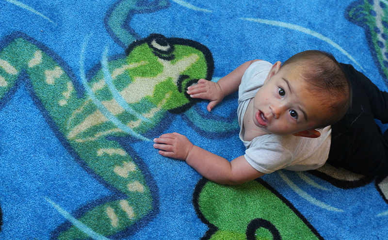 A baby on a rug. The rug has a stylized illustration of a frog partially submerged under water.