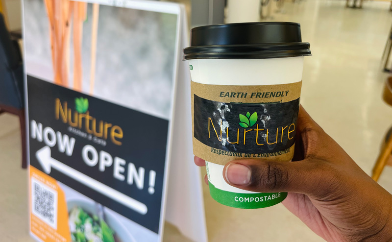 A person holding a coffee cup from Nurture next to a Nurture sandwich board style sign.