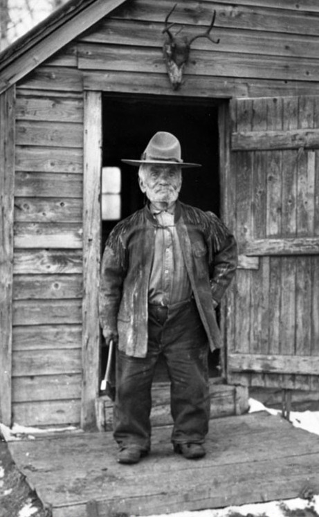 A short man standing in the doorway of a wooden cabin, dressed in Western attire.