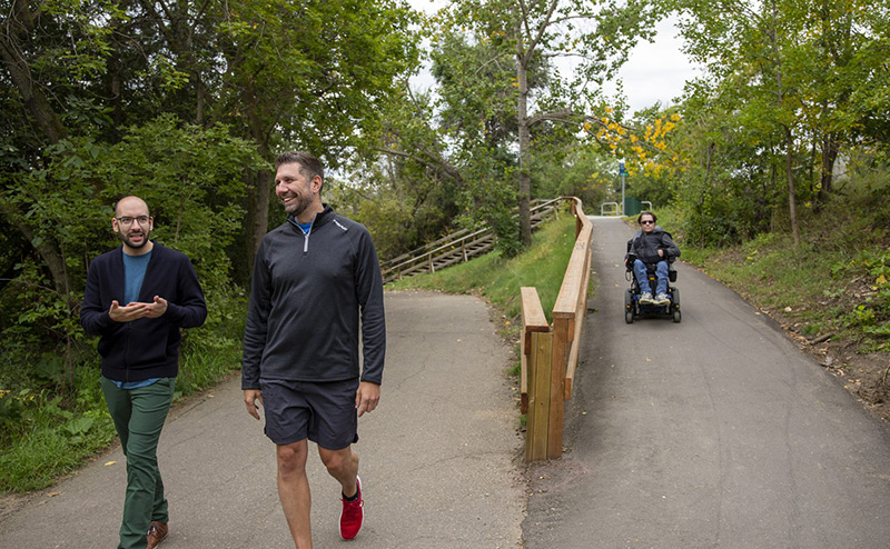 persons walking on trail beside an accessible pathway with person in a motorized wheelchair