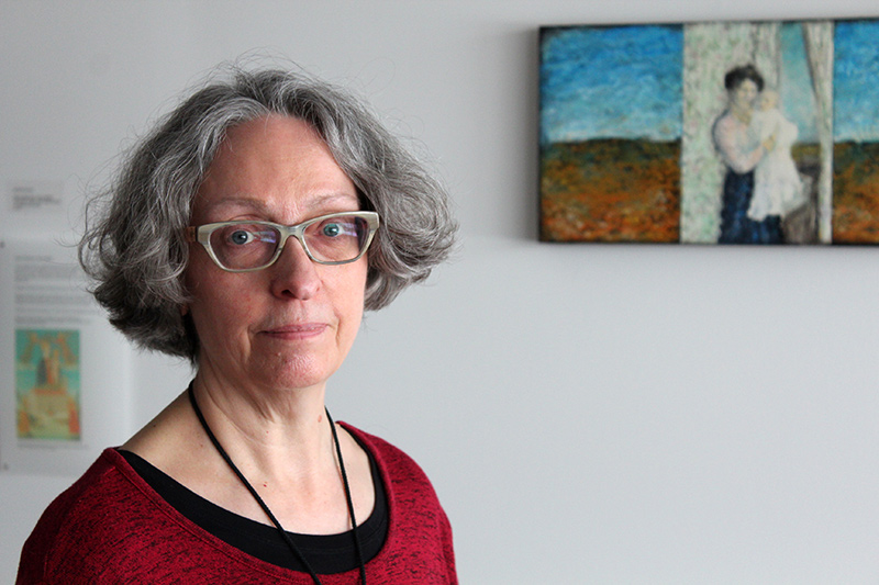 A woman with jaw-length grey hair and glasses, standing in front of a painting.