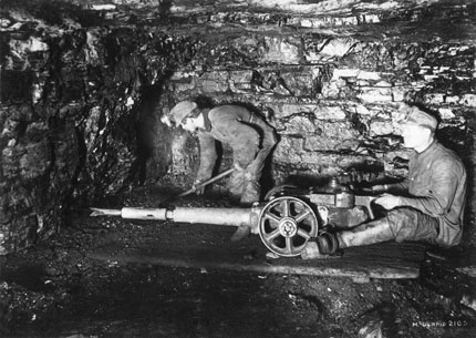 Two workers operating a punch miner. This machine looks like a long drill-like machine on wheels.