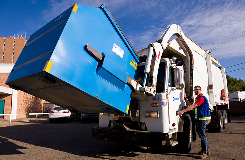 A Multi-family Collector standing next to a front-loading waste collection vehicle with a recycling bin on the forks.