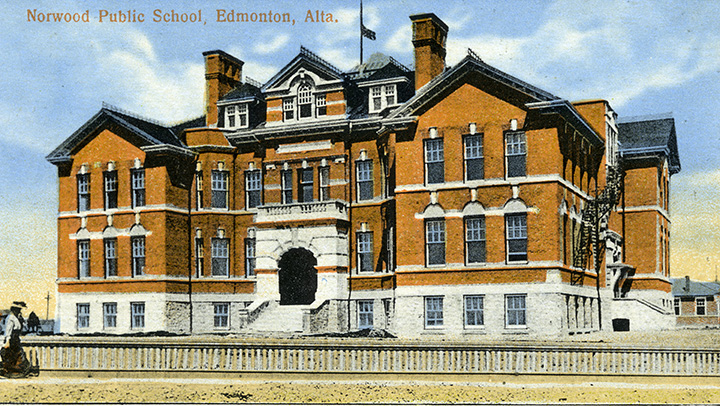 Colourized photo. Orange and white multi-story school building with a gray roof. A woman is walking past a fence in the foreground. Overlaid text at the top of the photo reads "Norwood Public School, Edmonton, Alta." A flag pole sits atop the school. The flag is at half mast, but the design is hard to make out.
