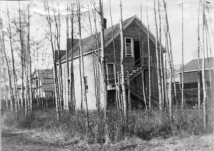 Black and white photo of a small two-story building surrounded by trees. The building has an outdoor staircase leading from the ground to the second story. Several other buildings can be seen nearby.
