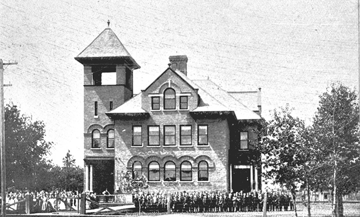 Black and white photo. Large multi-story building. A group of people, most likely students and teachers, stands outside. Several trees surround the building. There is a spiked fence in the foreground, near the bottom of the photo.