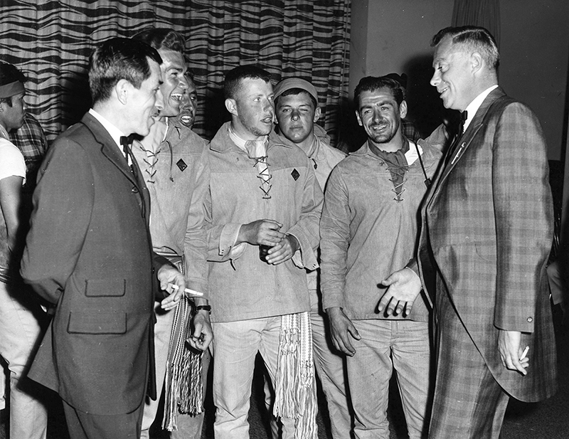 A black and white photograph of five men standing in a semicircle while talking and laughing.