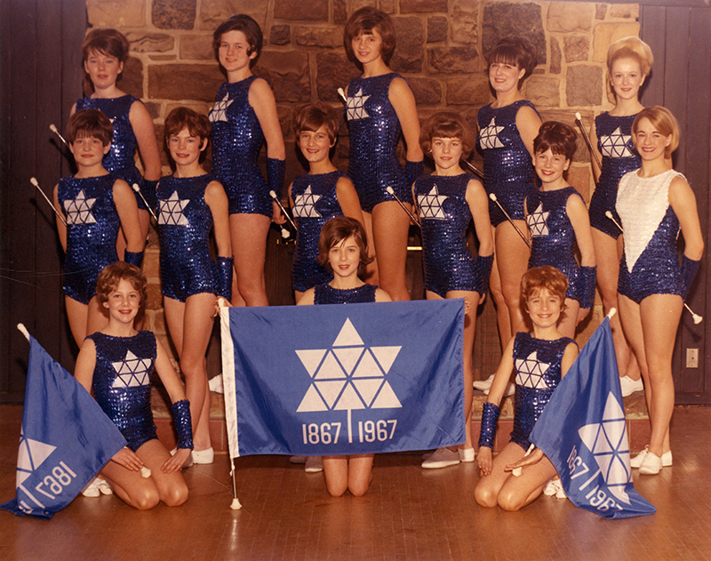 A group photo of girls dressed in blue Centennial gymnastics uniforms and holding centennial flags.