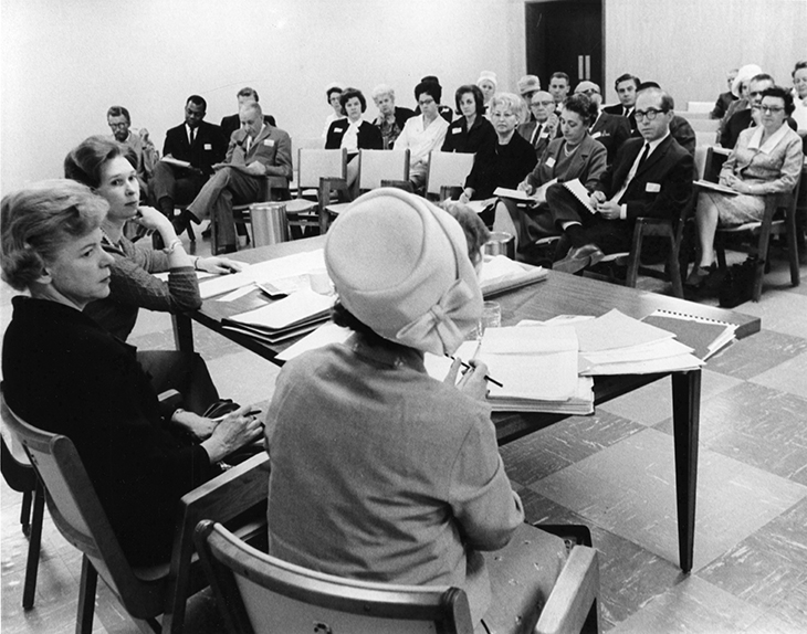 A black and white photograph of several people sitting in a room. They are seated to face three women at a head table.