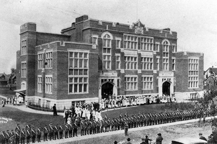 Black and white photo. A large multi-story school building. Each wall of the building has windows aligned in a grid. A large group of people, presumably teachers and students, stand along a path outside the building, leading up to the entrance. Another group of people, who appear to be soldiers, stand in a line along a road outside of the school building.