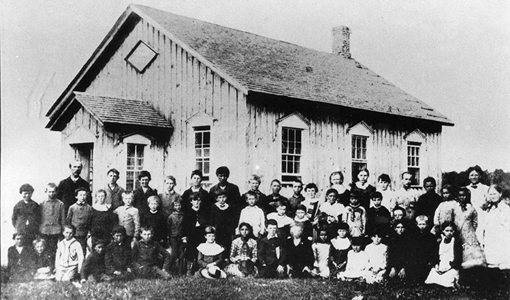 Black and white photo of a schoolhouse with a large group of children and some adults posing for a group photo in front of the building.