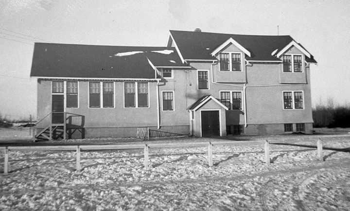 Black and white photo. A small house-shaped school building. A small shed-shaped structure in front of the building appears to be the front entrance. There also exists a smaller door on the left side of the building, with a set of wooden stairs leading up to it.