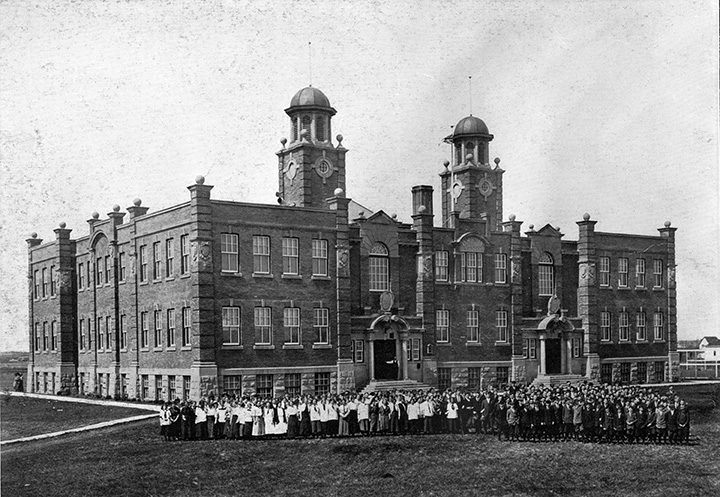 Black and white photo. A large multi-story school building. A large group of people, presumably students and teachers, stand outside.