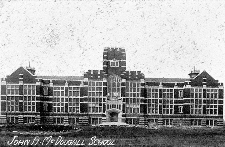 Grainy black and white photo. Front view of a large multi-story school building. Windows arranged in grids line the front of the building. A single entrance door sits at the centre, with a small flight of stairs leading up to it. Text overlaid on the bottom-left of the image reads "John A. McDougall School".