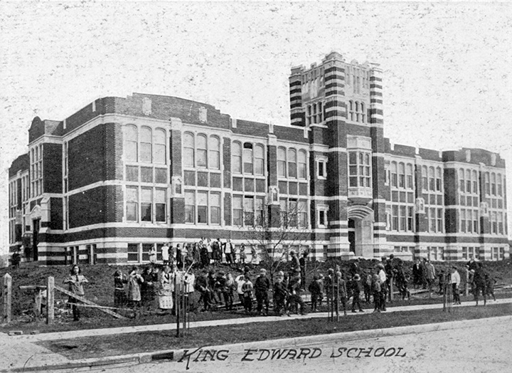 A brick school with several white windows, and students in front of it.