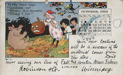 A postcard from October 1910. A drawing depicts a woman holding a jack-o-lantern. Two children follow behind her, and a bear sits in front of her, looking scared. A caption reads: "Teddy bear says, 'Goodness me! What's this dreadful thing I see!'" The post card has hand-written text which reads: "Your new costume will be a success if the material comes from this store. Don't miss seeing our line of Fall and Winter Dress Fabrics."