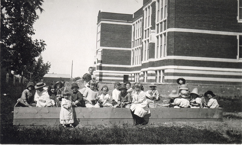 A black and white photograph featuring  children playing in a wooden sandbox beside a large brick school.