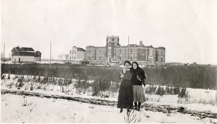 Black and white photo. Two women, each with an arm around the other, posing for a photo. Far in the background, Highlands School can be seen. Two other house-shaped buildings sit nearby.