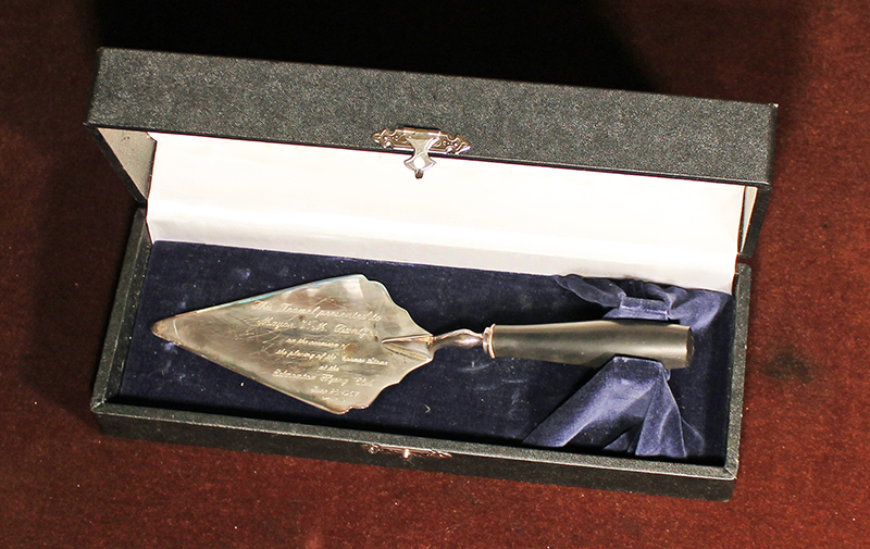 Colour photo of a trowel in a display case. The trowel is engraved with the text "This Trowel presented to Mayor V.M. Dantzer on the occasion of the placing of the Corner Stone at the Edmonton Flying Club June 22 1967"