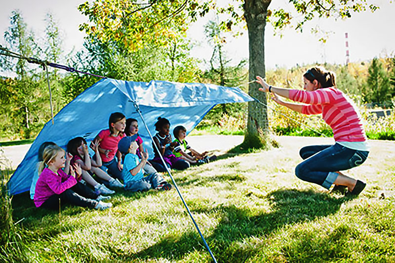 An instructor demonstrating to children. The children are in a lean-to shelter made from a blue tarp. The sun is very bright.