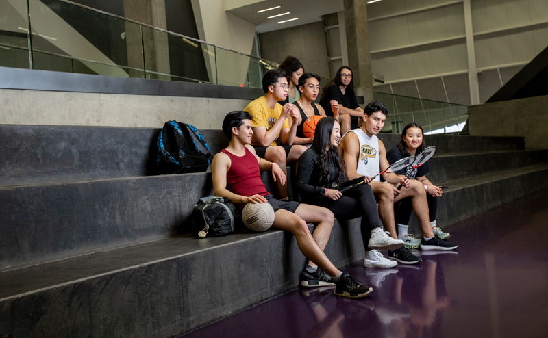 A group of teens with sporting equipment sitting in a rec centre.