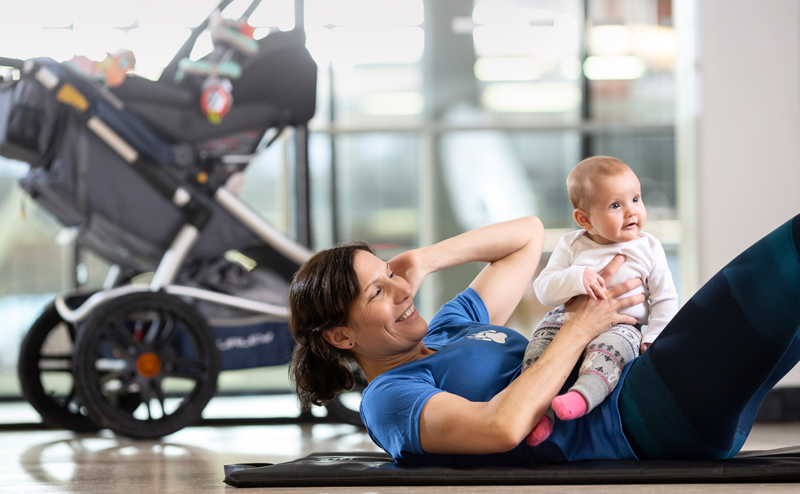 A woman laying on the floor, holding a baby while doing crunches. The baby's stroller is visible in the background.
