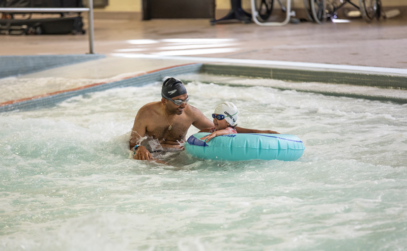 A man standing in a pool, holding a floatation device with a child in it. Both the man and child are wearing swimming caps and goggles.