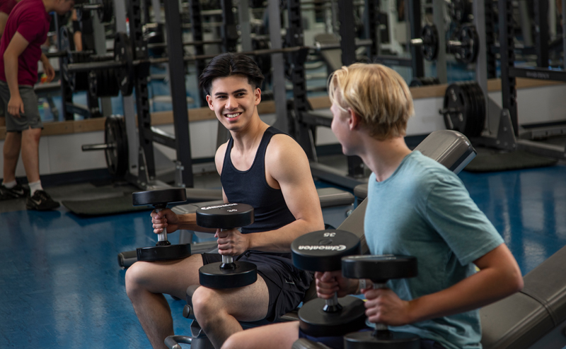 Two young men sitting on a bench holding dumbbells.