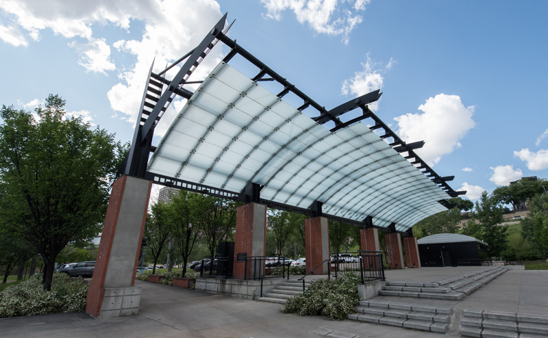 Photo of the Shumka Stage at Louise McKinney Riverfront Park. The stage is a raised platform with steps, covered by a translucent awning overhead.