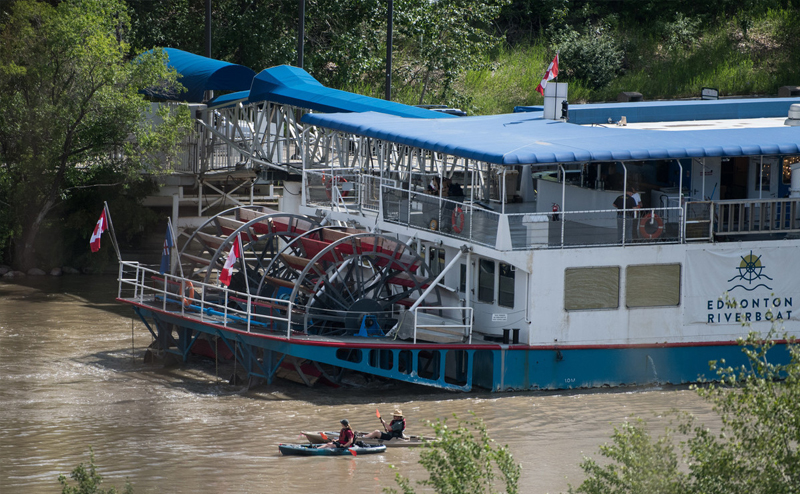 Edmonton Riverboat moving down a river with two people in canoes beside it.
