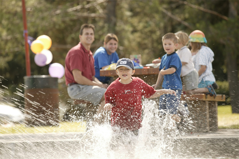 A child playing in a spray park as his family watches from a picnic table.