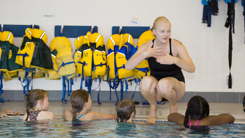 An instructor, squatting at the edge of a pool, talking to a group of kids in the pool.