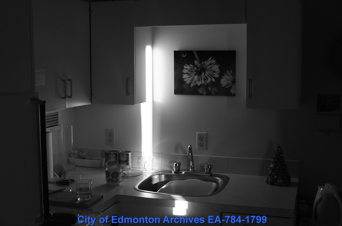 A black and white photo of a kitchen sink, illuminated by a long bright light. 