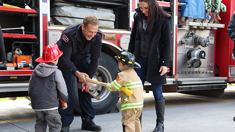 An adult and two kids dressed as firefighters meet a firefighter. The firefighter hands one of the kids a fire axe.