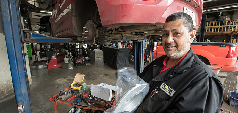A man working in an auto garage. A car with no wheels sits on a lift behind him.
