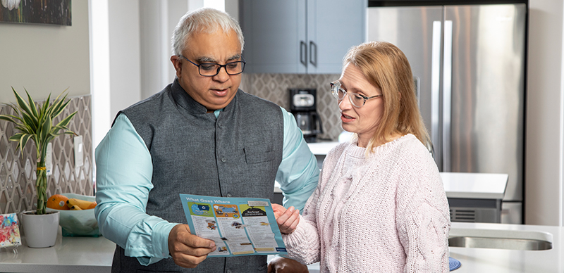 A man and woman standing in a kitchen reviewing a "What goes where" waste document