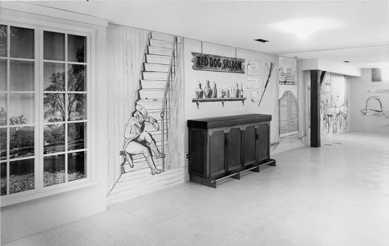 A black and white photo of a basement painted with a western theme. Paintings on the wall include a cowboy sleeping on some stairs (the stairs are also painted on the wall), saloon doors, and a wooden sign that says "Red Dog Saloon". Below the painted sign is a painting of a shelf full of liquor.