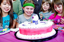 A group of four young girls excitedly looking at a birthday cake. The cake and table cloth are pink.