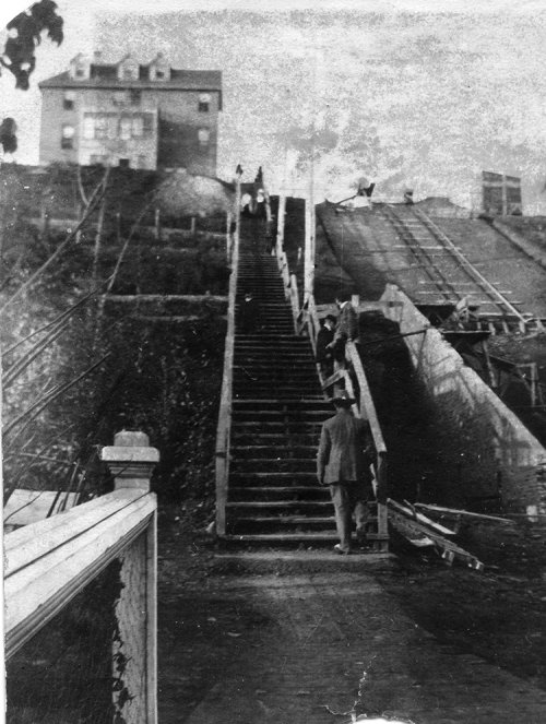 A black and white photograph of wooden stairs leading up a hill, alongside the early stages of a railway under construction.