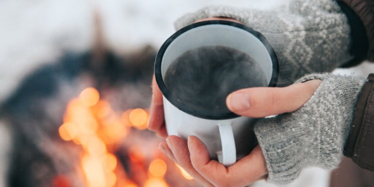 A person's hands in fingerless mittens holding a steaming mug near a campfire.