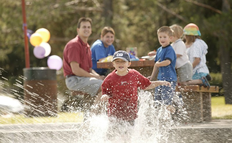 A child playing in a spray park as his family watches from a picnic table.