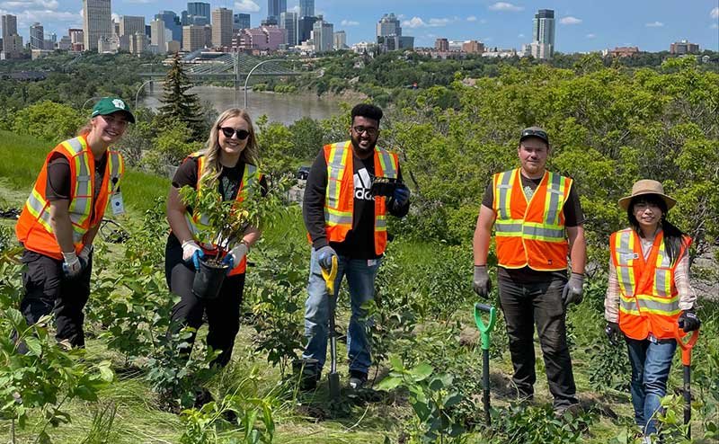 5 people wearing flourescent orange and yellow vests posing in a line with the trees they are about to plant 