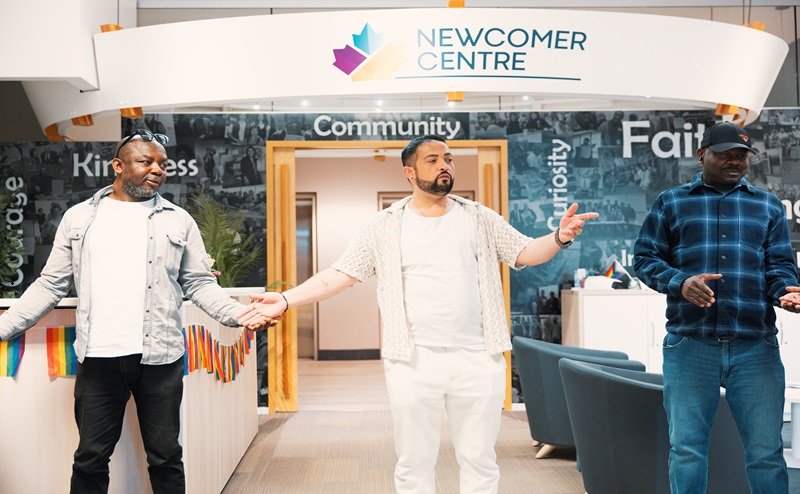 Three individuals holding hands in front of a "Newcomer Centre" sign.
